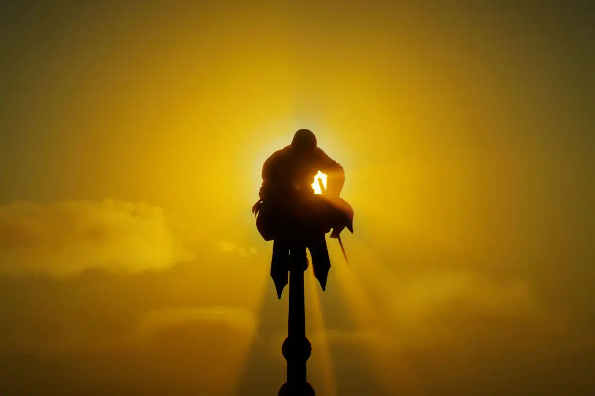 A cinematic image screenshot taken in Assassin's Creed Mirage, Basim Ibn Ishaq / Loki can be seen perched on top of a tower ready to do a Leap of Faith in front of the beautiful setting sun, god rays can be seen around Basim as they are of an Aura